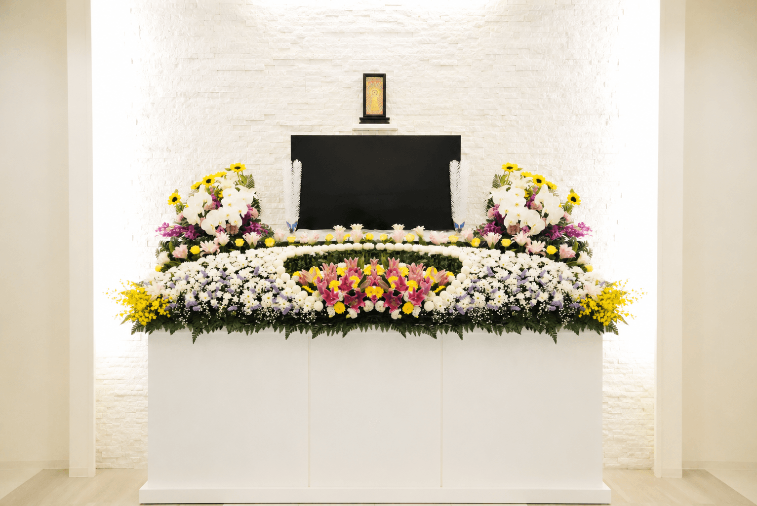 白い式場に組まれた、胡蝶蘭とひまわり・白菊による二段の生花祭壇